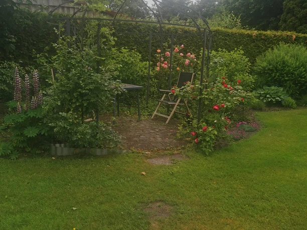 Sitzecke Privatgarten Hienen_Foto Hienen.jpg Eine gepflasterte Sitzecke unter einem Rosenbogen mit Stuhl und Tisch in einem üppigen, grünen Garten.