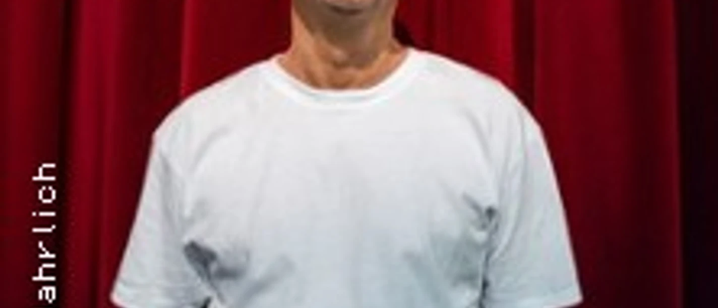 Eine lächelnde Person mit Glatze im weißen T‑Shirt vor geschlossenem roten Vorhang