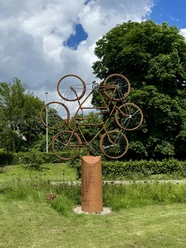 Eine Skulptur aus mehreren rostfarbenen Fahrrädern steht auf einer Wiese vor grünen Bäumen.