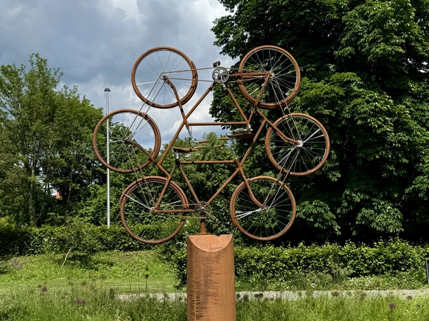 Eine Skulptur aus mehreren rostfarbenen Fahrrädern steht auf einer Wiese vor grünen Bäumen.