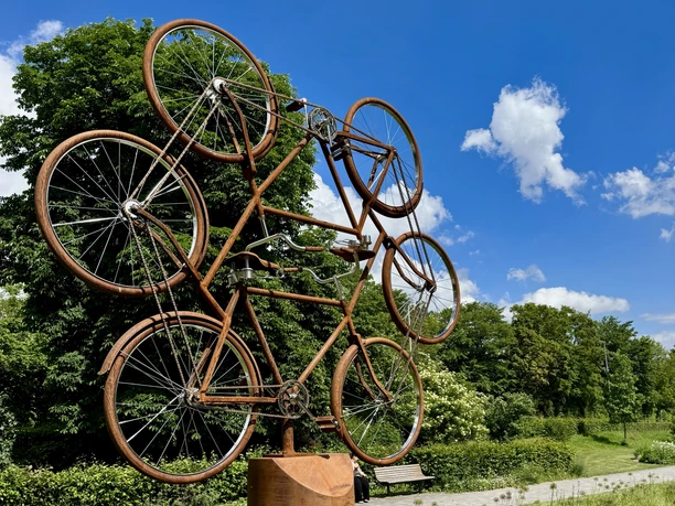 Kunstskulptur aus rostfarbenen Fahrrädern auf einem Sockel in einer grünen Parkanlage.