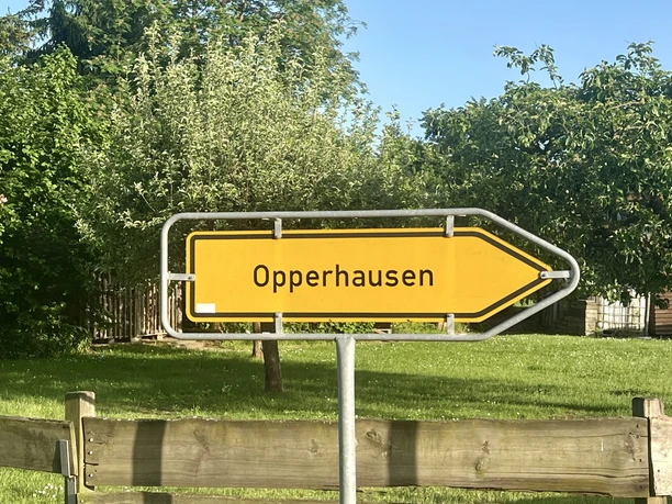 Schild Richtung Opperhausen, Hochformat Gelber Wegweiser nach Opperhausen vor einem Holzzaun, einer Wiese und Bäumen bei Sonnenschein.