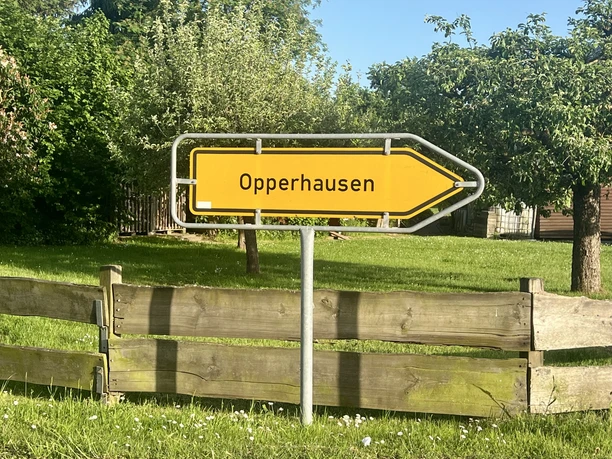 Schild Richtung Opperhausen, Querformat Gelber Wegweiser mit Pfeil nach rechts und der Aufschrift Opperhausen vor einem Holzzaun im Grünen.