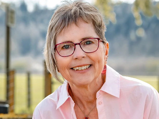 Porträt von Claudia Thienenkamp Lächelnde Frau mit Brille, pinkem Hemd, Armbändern und Uhr, stützt sich auf Holzgeländer draußen.