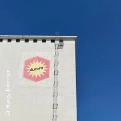 Weiße Hausfassade mit Aurora-Logo, Leiter und blauem Himmel am Deutzer Hafen
