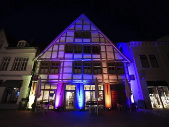 Ein Fachwerkhaus bei Nacht, beleuchtet in blau, pink und orange, mit offenen Sonnenschirmen davor.