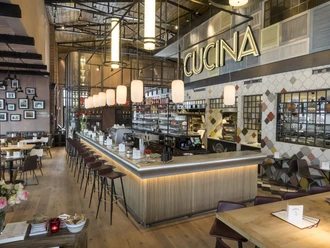 Barhocker stehen an einer großen Theke, in der sich Gläser, Tassen, eine Kaffeemaschine und ein Regal mit weiteren Gläsern und Flaschen befinden. Über der Theke hängt die Leuchtschrift "CUCINA" und davor mehrere Tischgruppen. Von der Decke hängen Lampen herab.