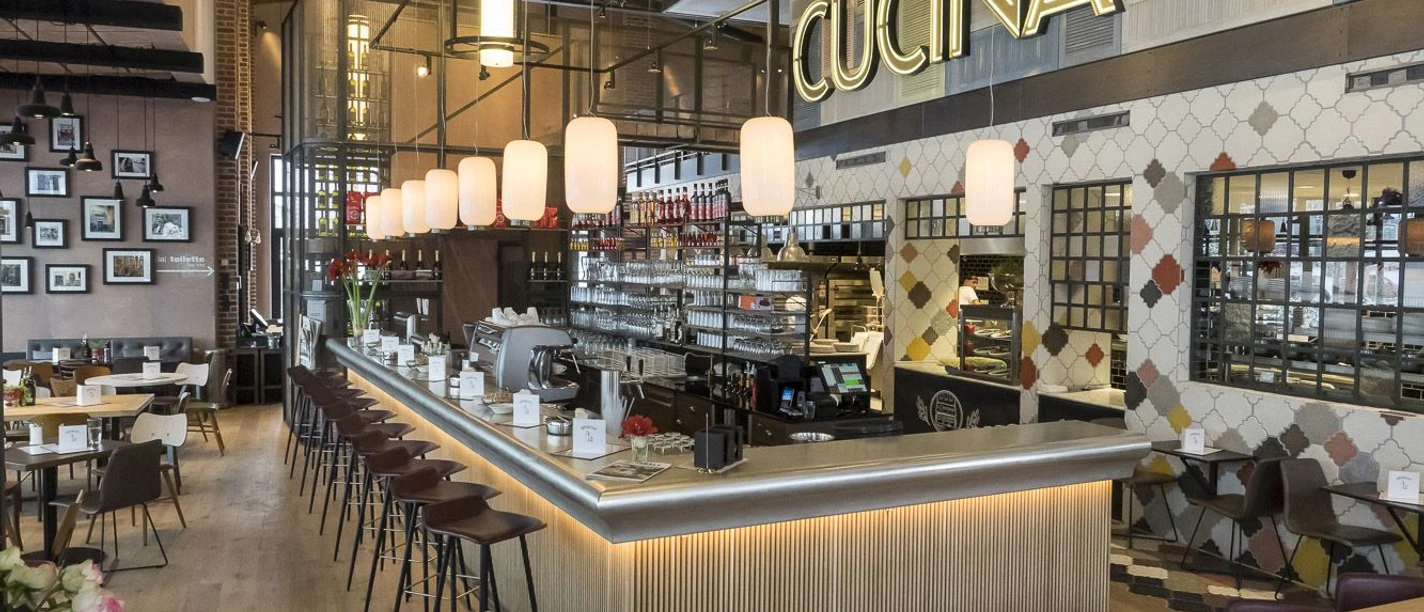 Barhocker stehen an einer großen Theke, in der sich Gläser, Tassen, eine Kaffeemaschine und ein Regal mit weiteren Gläsern und Flaschen befinden. Über der Theke hängt die Leuchtschrift "CUCINA" und davor mehrere Tischgruppen. Von der Decke hängen Lampen herab.