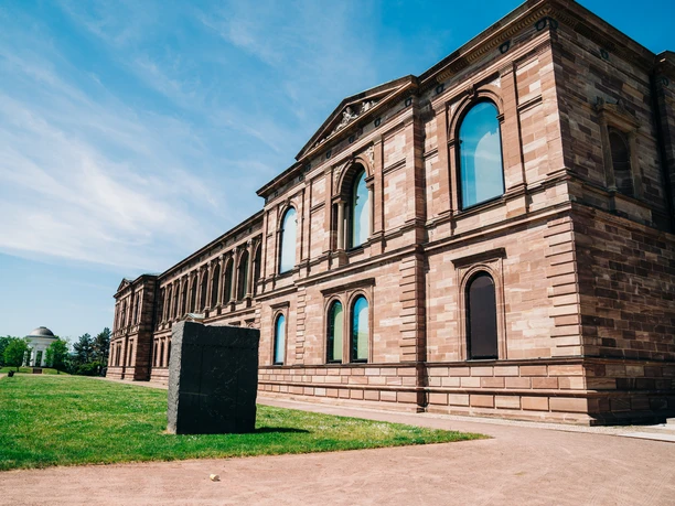 Großes, historisches Museumsgebäude aus rötlichem Sandstein mit reich verzierter Fassade und bogenförmigen Fenstern. Vor dem Gebäude erstreckt sich eine Grünfläche mit einem freistehenden, dunklen Steinblock im Vordergrund sowie einem hellen Kiesweg.