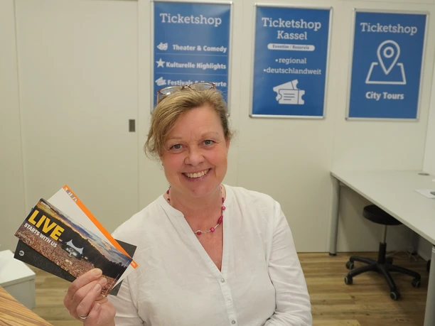 Eine Mitarbeiterin der Tourist-Information in Kassel präsentiert Tickets die in der Tourist-Information erworben werden können. Im Hintergrund sind blaue Plakate mit der Aufschrift "Ticketshop".