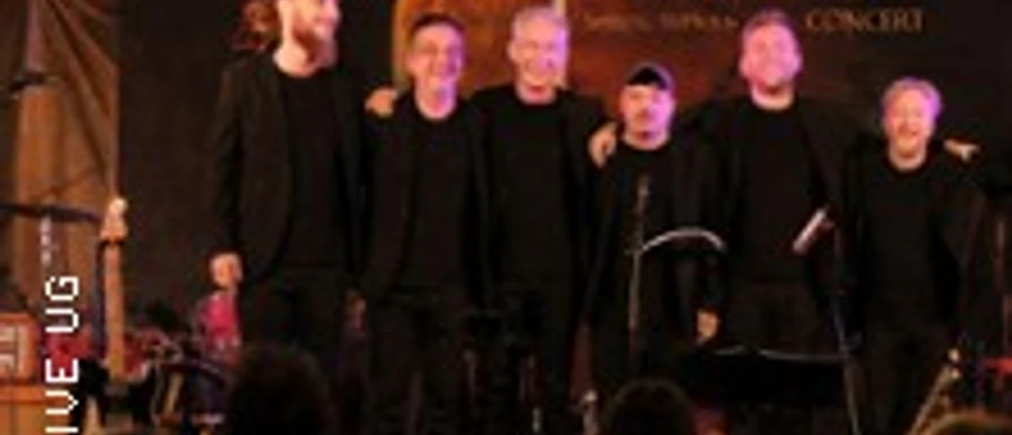 Mehrköpfige Musikgruppe in dunkler Kleidung auf beleuchteter Bühne vor Publikum