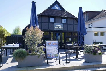 Moto-Kitchen-Bad-Zwischenahn-Aussenansicht.jpg Außenansicht des Restaurants Moto Kitchen in Bad Zwischenahn mit Terrasse und großen Sonnenschirmen.