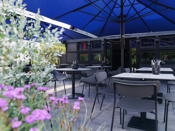 Moto-Kitchen-Bad-Zwischenahn-Terrasse.jpg Die moderne Terrasse des Restaurants Moto Kitchen mit grauen Tischen unter blauen Sonnenschirmen.