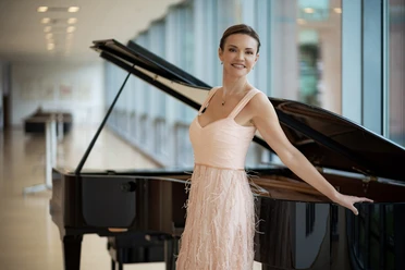 Olga Andryushchenko Eine lächelnde Frau im Abendkleid lehnt an einem Flügel in hellem Saal mit großen Fenstern.