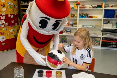 Kinderhaus WM Nachmittag Ein Mädchen im Deutschland-Trikot bemalt mit dem Maskottchen Lükko einen Fußball in Schwarz-Rot-Gold.