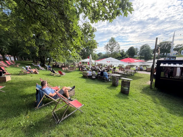 Weserstrand Beach Nienburg Gäste entspannen in Liegestühlen auf einer Wiese am Weserufer unter großen, schattigen Bäumen.
