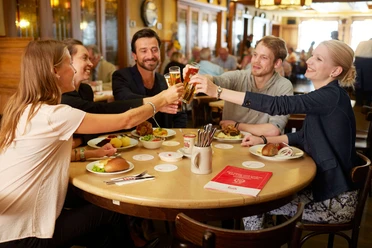 Früh am Dom A group of people are sitting around a round table, clinking glasses of Kölsch beer and having small Cologne dishes in front of them.