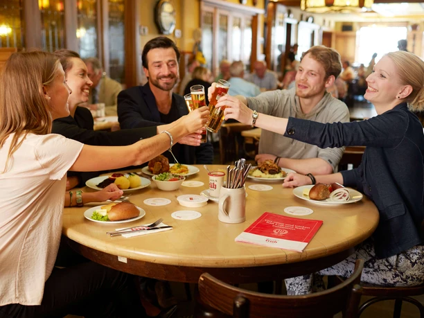 Früh am Dom A group of people are sitting around a round table, clinking glasses of Kölsch beer and having small Cologne dishes in front of them.