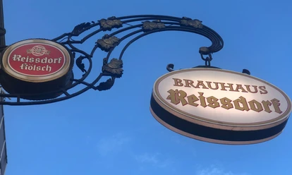 Zwei kunstvoll verzierte Brauhaus-Schilder, "Reissdorf Kölsch" und "Brauhaus Reissdorf", vor blauem Himmel.
