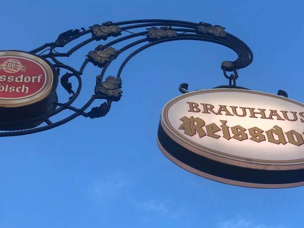 Reissdorf Zwei kunstvoll verzierte Brauhaus-Schilder, "Reissdorf Kölsch" und "Brauhaus Reissdorf", vor blauem Himmel.