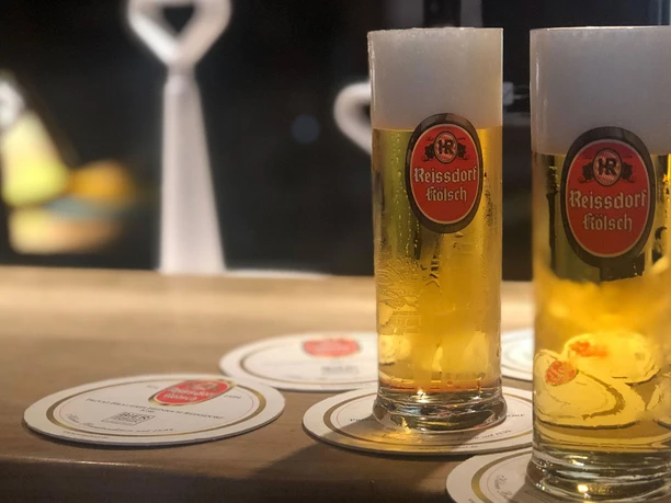 Reissdorf Zwei Reissdorf-Kölsch-Gläser auf einem Tresen, umgeben von Bierdeckeln, in einem gemütlichen Ambiente.