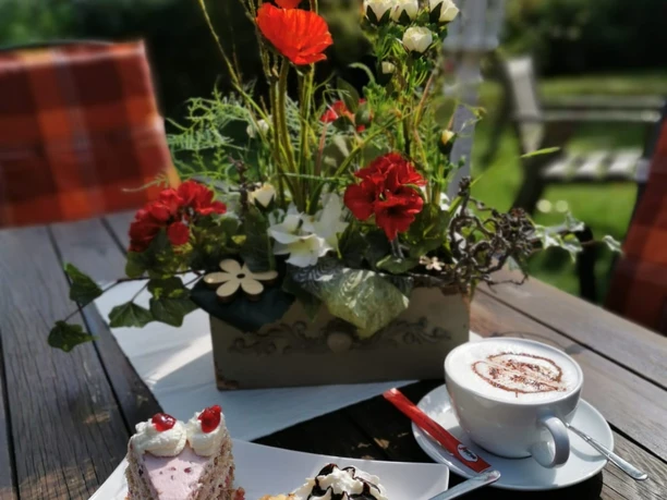 Floristik unterm Storchennest Blumenarrangement mit roten Mohnblumen, daneben Tortenstücke und eine Tasse Cappuccino auf Holztisch.