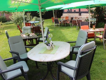 Floristik unterm Storchennest Ein Gartenbereich mit runden Tischen und Stühlen unter grünen Sonnenschirmen, umgeben von Pflanzen.