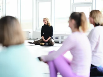 Eine Yoga-Lehrerin sitzt lachend auf einer Matte vor den Teilnehmenden in einem hellen Raum.