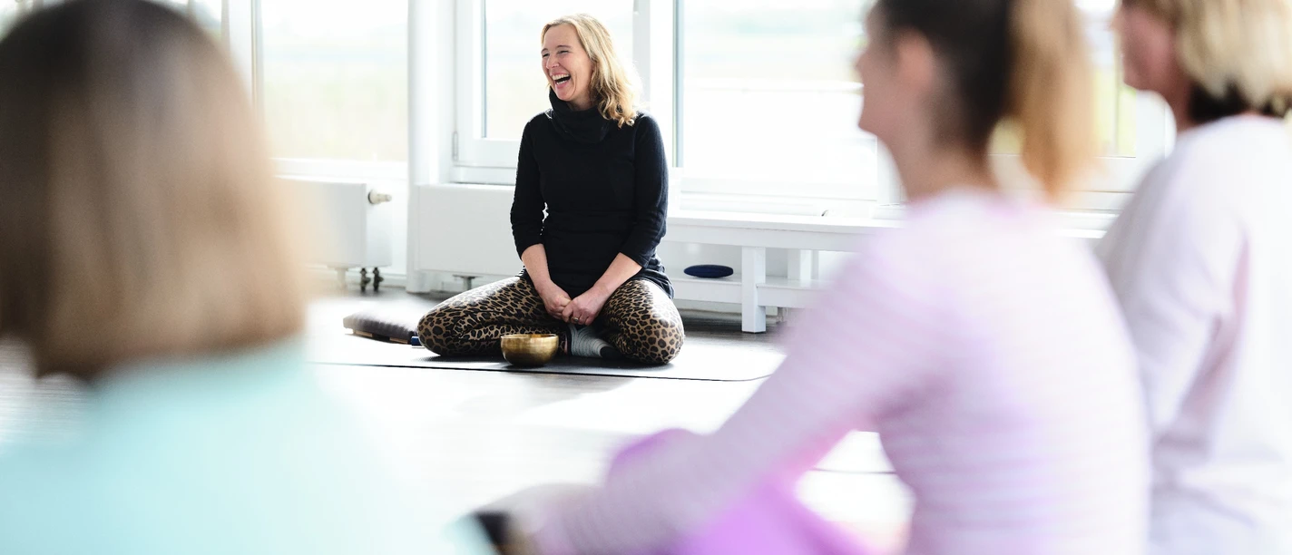 Yogalehrerin Wiebke Niebel sitzt lachend im Schneidersitz in hellem Raum.jpg Eine Yoga-Lehrerin sitzt lachend auf einer Matte vor den Teilnehmenden in einem hellen Raum.