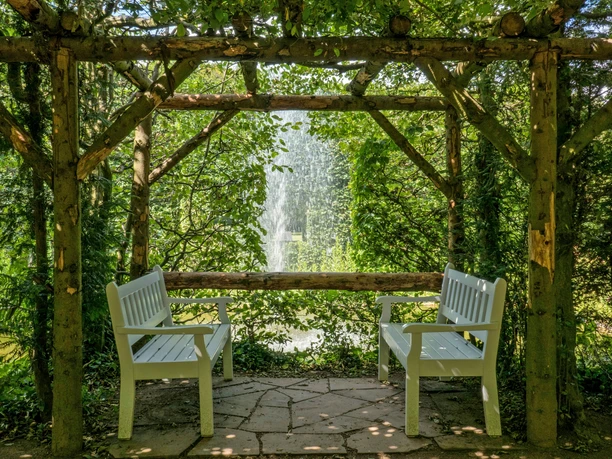 Heckengarten Weiße Bänke unter Holzpergola mit Blick auf Springbrunnen im Heckengarten