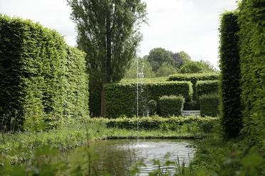 Heckengarten mit Teich, Springbrunnen und weißer Bank zwischen hohen Hecken