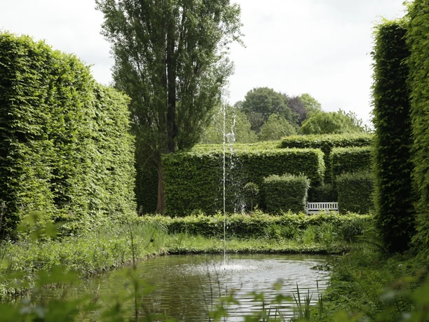 Heckengarten mit Teich, Springbrunnen und weißer Bank zwischen hohen Hecken