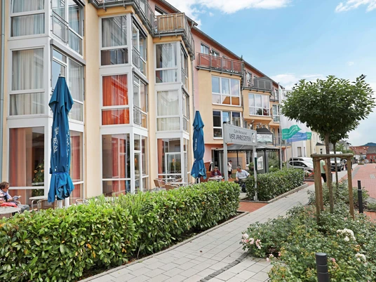 carpe-diem_ambulante-pflege-juli-2018-14 Mehrstöckiges Gebäudekomplex mit Balkonen, großen Fenstern und Café, umgeben von einem Gehweg und Grünflächen.