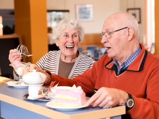 cafeteria_1-6 Seniorenpaar genießt Kaffee und Kuchen in einer modernen Cafeteria, umgeben von farbenfroher Dekoration.