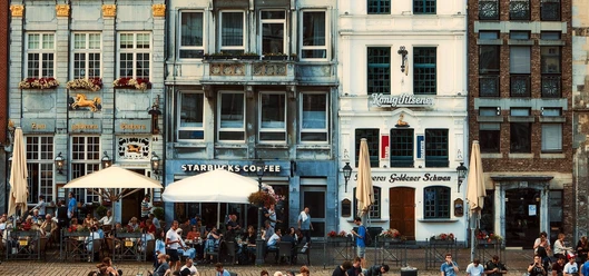Vier schmale, mindestens dreistöckige Häuser stehen aneinandergereiht auf dem Marktplatz in Aachen. In den Erdgeschossen befinden sich Gastronomien, wovon eine das Restaurant Goldener Schwan ist. Vor den Lokalen stehen Sitzgruppen und Schirme.