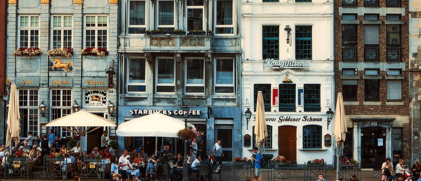 Markt und Goldener Schwan (c) Pixabay Vier schmale, mindestens dreistöckige Häuser stehen aneinandergereiht auf dem Marktplatz in Aachen. In den Erdgeschossen befinden sich Gastronomien, wovon eine das Restaurant Goldener Schwan ist. Vor den Lokalen stehen Sitzgruppen und Schirme.