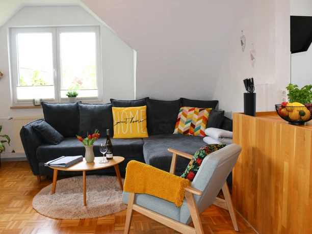 Ferienwohnung-Familie-Baasch-Wohnzimmer.jpg Wohnzimmer mit dunkler Eckcouch, farbigen Kissen, Sessel und einem Holztresen.