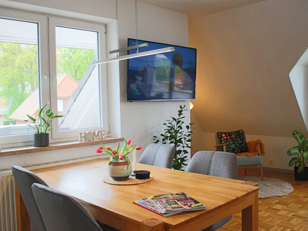 Heller Essbereich der Ferienwohnung mit Holztisch, grauen Stühlen und Blick ins Grüne.