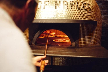 gold-of-naples Ein Mann schiebt eine Pizza auf einer flachen Schaufel in einen großen Pizzaofen, in dem sich bereits weitere Pizzen befinden.