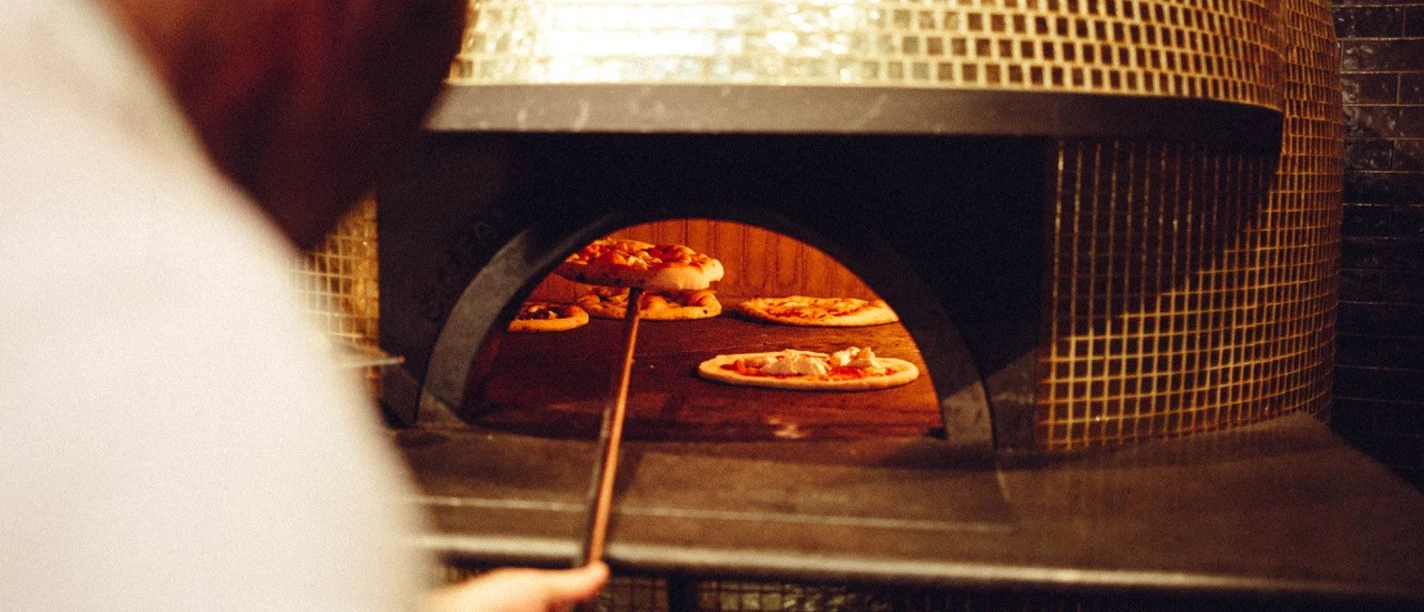 gold-of-naples Ein Mann schiebt eine Pizza auf einer flachen Schaufel in einen großen Pizzaofen, in dem sich bereits weitere Pizzen befinden.