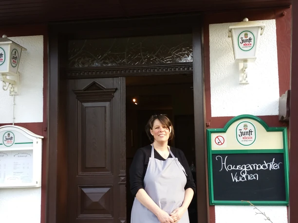 Eingang Eine Frau in Schürze steht lächelnd vor dem Eingang eines Cafés mit Holzdekoration.