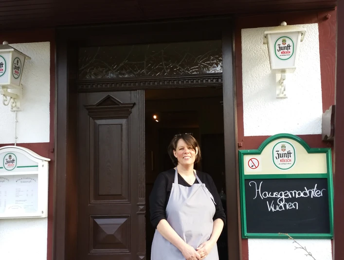 Eingang Eine Frau in Schürze steht lächelnd vor dem Eingang eines Cafés mit Holzdekoration.