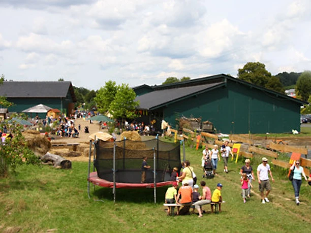 Außengelände Familienfreundlicher Bauernhof mit Spielplatz, grünen Scheunen und vielen Besuchern bei strahlendem Wetter.