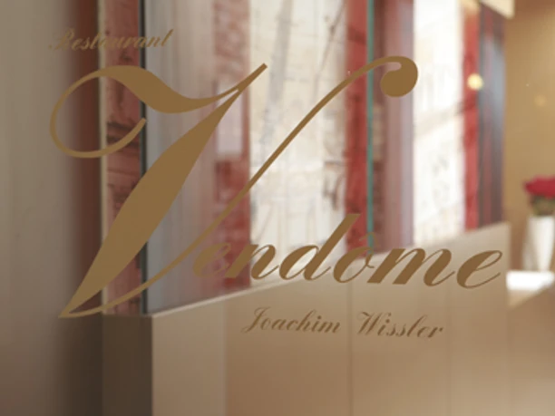 Restaurant Vendome Logo Schriftzug "Vendome" mit eleganter Schrift auf einer Glasfront, dahinter unscharfe Innenraumszene.