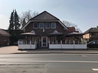 schlemmer-grill-haren Backsteinhaus mit zwei überdachten Terrassenbereichen und weißen Geländern an einer Straße.