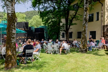 Erzquell Brauerei Sommerfest Menschen sitzen auf Wiesenstühlen beim Brauereifest zwischen Bäumen und gestapelten Getränkekisten.
