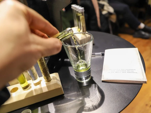 Absinth-Tasting in Köln Hand gießt Absinth aus einem Röhrchen in ein Glas auf einem Tisch mit Unterlagen