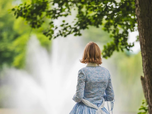 Geschichte im Grünen Rückansicht einer Frau in einem blauen, geblümten Kleid im Park mit Blick auf einen Springbrunnen.