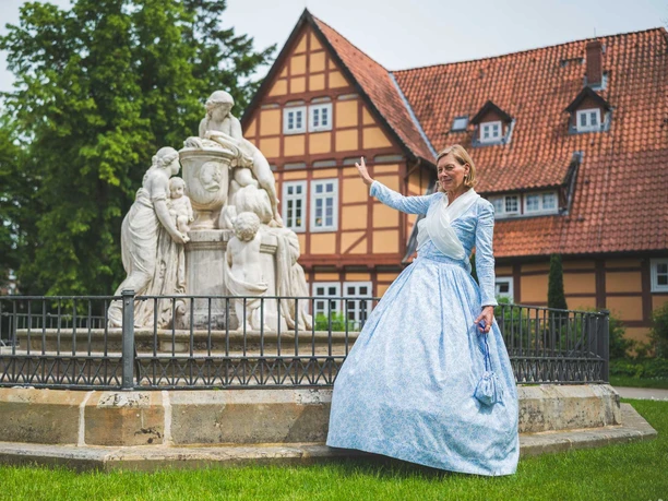 Geschichte im Grünen Eine Gästeführerin in historischem Kleid winkt vor einem Brunnen im Französischen Garten in Celle.