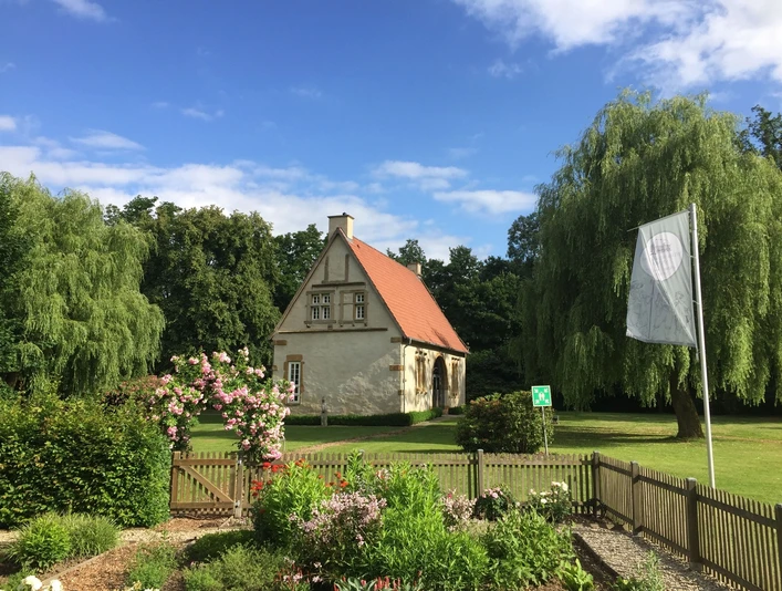 Torhaus einer historischen Burganlage mit rotem Giebeldach, umgeben von Garten und Fahne.